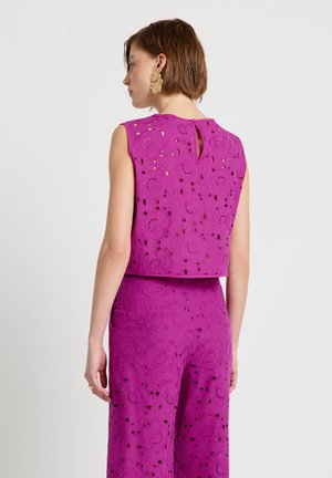 Donna che indossa una canottiera smanicata magenta con motivo floreale traforato e pantaloni a vita alta abbinati, vista di lato, con capelli corti castani e grandi orecchini d'oro.