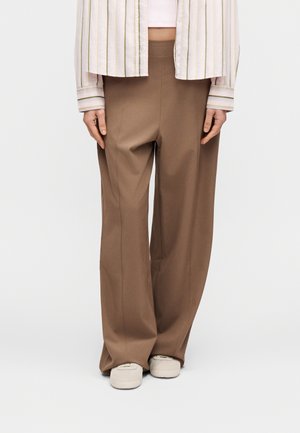 Pruunid laiad püksid, millel on sile pind, õmmeldud õmblused ja lõdvestunud lõige. Sobitatud heledate jalanõude ja triibulise särgiga.