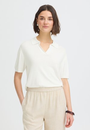 Moteris su trumpais rudais plaukais, vilkinti baltą trumpomis rankovėmis su apykakle marškinius ir smėlio spalvos kelnės su elastine juosmens juosta, stovinti prie vienspalvio fono.