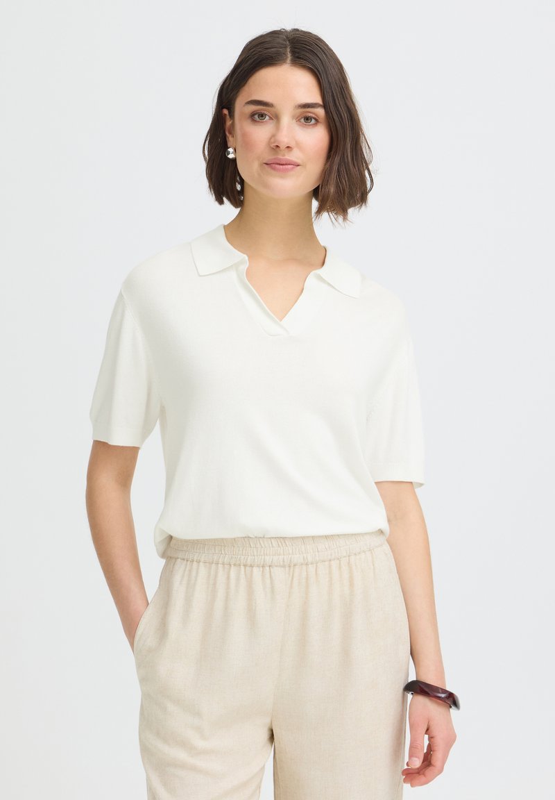 Femme aux cheveux courts et bruns portant une chemise blanche à manches courtes avec col et un pantalon beige à taille élastique, debout devant un fond uni.