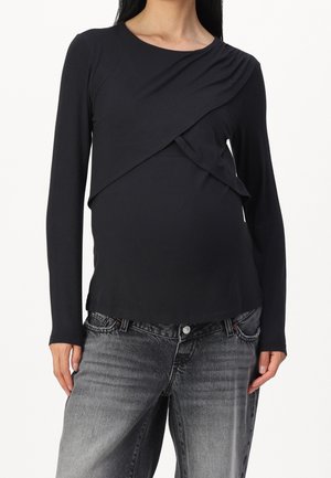 Kobieta w czarnym długim topie z warstwowym przodem i spranych czarnych dżinsach na białym tle.