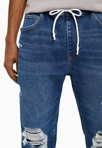 Denim joggers in medium blue featuring frayed knee patches, a drawstring waistband, and a relaxed fit with side pockets.