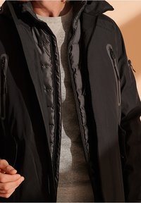 Chaqueta negra resistente al clima con capucha, bolsillos con cremallera, forro acolchado y exterior liso. Capa interior en gris. Diseño minimalista.