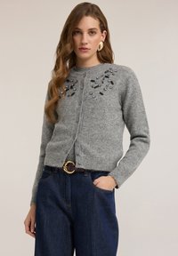 Šedý vlněný cardigan s dekoračními ozdobami u výstřihu, zapínáním na knoflíky a střihovým designem, ve spojení s tmavě modrými džínami.