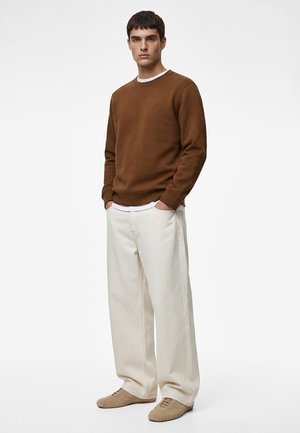 Junger Mann mit braunem Sweatshirt, weißen Hosen mit weitem Bein und beigen Schuhen, stehend mit den Händen in den Taschen vor einem einfarbigen Hintergrund.