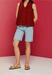 Maroon bezrękawnik z guzikami, w zestawieniu z jasnoniebieskimi szortami dżinsowymi i beżowymi klapkami na różowym tle.
