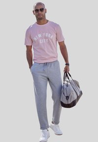 Helles pinkes T-Shirt mit dem Text "NEW YORK CITY", graue Sweatpants, weiße Turnschuhe und eine grau-braune Sporttasche. Sonnenbrille getragen.