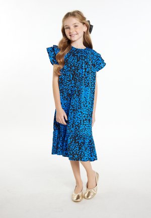 Blaue Leopardenmuster-Kleid mit kurzen Rüschensäumen und einem gestuften Saum. Getragen mit goldenen Ballerinas. Hintergrund ist weiß.