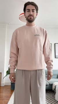 Svetlo roza sweatshirt z okroglim izrezom in logotipom; v kombinaciji s svetlobnimi širokimi hlačami. Mehak material, sproščen kroj. Notranji prostor z rastlinami.