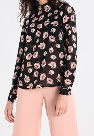 Woman wearing a black blouse with pink floral print and light pink wide-leg pants, standing with one hand relaxed by her side.