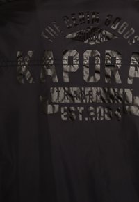 Black fabric featuring embossed text in varying opacities, including "KAPORA" and "EST.2004," with a smooth texture.