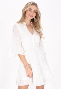 Balta suknelė su plačiomis rankovėmis, siuvinėtu gėlių raštu, V formos iškirpte ir sluoksniuotu sijonu. Papuošalai apima sluoksniuotą apyrankę ir žiedus.