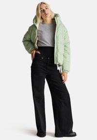 Giacca a puffo verde chiaro con cappuccio, polsini elasticizzati e zip; camicia grigia sotto; pantaloni neri a gamba larga; superfici lisce in tutto.