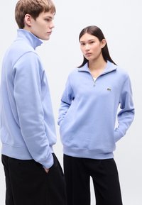 Ανοιχτό γαλάζιο φούτερ με φερμουάρ, με ριμπ λαιμόκοψη και μανσέτες, με μικρό πράσινο λογότυπο στο στήθος. Συνδυάζεται με μαύρο παντελόνι.