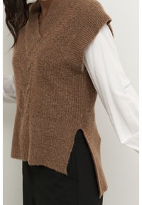 Vrouw draagt een bruine gebreide mouwloze trui over een wit shirt met lange mouwen en zwarte broek, zijaanzicht.