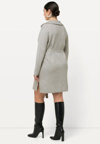 Cappotto grigio con cintura e maniche lunghe, caratterizzato da un colletto classico, fessure laterali e un tessuto testurizzato; abbinato a stivali neri sopra al ginocchio.
