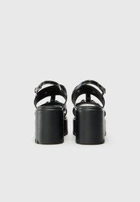 Black platform sandals with wide straps, a smooth finish, and buckle closures, featuring a chunky heel and rounded toe design.