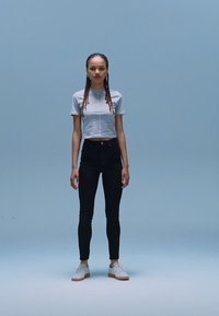 Balts īss krekls, melnas augstās jostas bikses un balti sporta apavi ar gumijas zolēm. Vienkāršs fons, modelis stāv taisni, neitrālā pozā.