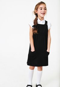 Czarna sukienka z okrągłym dekoltem, bocznymi kieszeniami i guzikami. Noszona na białej koszuli z kołnierzykiem. Białe skarpetki do kolan i czarne buty.