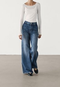 Jeans blu a gamba larga con texture sbiadita e vita alta, abbinati a un top aderente bianco a maniche lunghe. Scarpe basse nere completano il look.