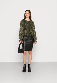 Blusa verde transparente com estampado de leopardos, mangas elásticas, saia lápis preta em couro, mala preta trançada e botas pretas à altura do tornozelo com detalhes em metal.