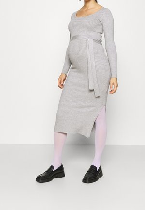 Gravid person iført en ribbet, langærmet, grå strikket kjole med bælte, lyserøde strømpebukser og sorte loafers, stående foran en hvid baggrund.