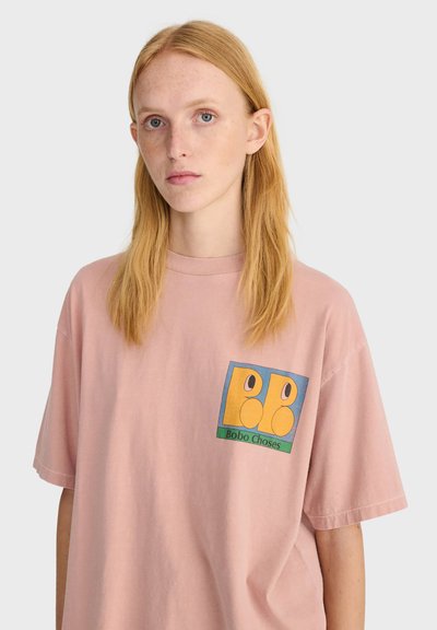 Jeune femme aux longs cheveux roux portant un t-shirt ample rose clair avec un logo coloré "Bobo Choses" sur la poitrine.