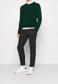 Vihreä neulottu villapaita, jossa kirjailtu logo, valkoinen kauluksellinen paita alla, yhdistetty tummiin ruudullisiin housuihin ja valkoisiin tennareihin.