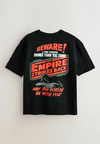 Cămașă T-shirt din bumbac negru, cu un imprimeu grafic ce include text în roșu și alb. Designul conține „Atenție!” și „Împăratul loveste din nou”, alături de imaginea unui personaj.