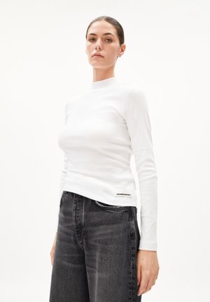 Weißes, geripptes Langarmshirt mit hohem Ausschnitt. Kombiniert mit dunkelgrauen Jeans aus Denim, die ein Etikett am Saum haben.
