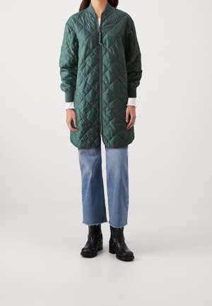 Femme portant un long manteau matelassé vert foncé avec fermeture éclair, un jean court bleu et des bottines noires à talons sur fond blanc.