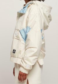 Chaqueta blanca y azul claro con capucha, que presenta un diseño a bloques de color, puños elásticos y un parche de logo en la manga. Tejido texturizado.