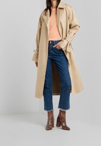 Beige trenchcoat över en orange tröja, kombinerad med blå jeans och bruna ankelboots med ormskinnsmönster. Har bred krage och ärmuppslag med bälte.