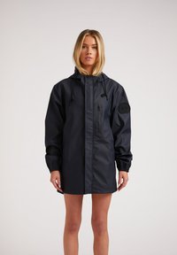 Veste imperméable navy avec capuche, poche zippée, poignets élastiques et un écusson logo sur la manche. Texture lisse et soyeuse avec un design jusqu'aux genoux.