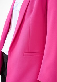 En ljusrosa blazer som bärs över en vit mönstrad skjorta och svarta byxor, som visar blazerns framficka och reversdetalj.