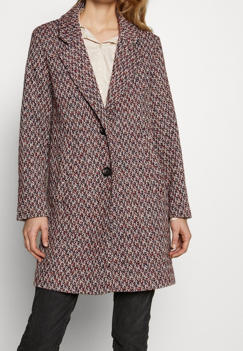 Femme portant un manteau en chevrons multicolore texturé, mi-long, à deux boutons, par-dessus un chemisier clair et un pantalon foncé.