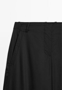 Pantalons noirs exposés, avec une taille, des détails de plis et des poches latérales. Le tissu semble lisse et léger.