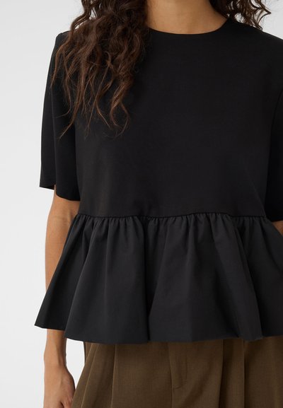 Femme portant un haut noir à manches courtes avec une taille peplum froncée et un pantalon marron, montrée du cou jusqu'à mi-cuisse.