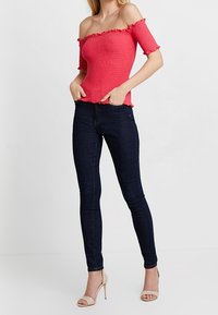 Off-shoulder pink top med smocket tekstur, rynkede kanter, kombineret med mørke skinny jeans og beige højhælede sandaler.