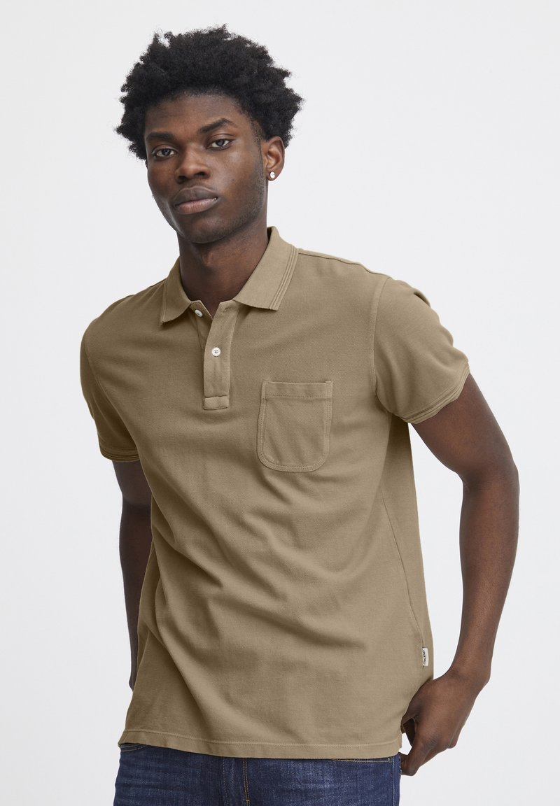 Camisa polo caqui de manga corta, con collar y un bolsillo en el pecho. Hecha de algodón, presenta detalles de costura sutiles y una etiqueta de logo en el lateral.