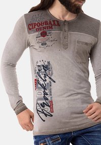 Camisa beige de manga larga con hombros grises contrastantes, con texto y gráficos impresos, tapeta de tres botones y bolsillo en el pecho.
