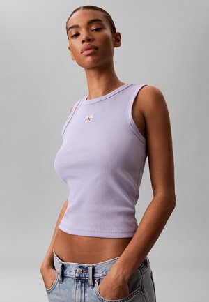 Młoda kobieta nosząca dopasowany lawendowy top bez rękawów i jasnoniebieskie dżinsy, pozująca z rękami w kieszeniach na szarym tle.