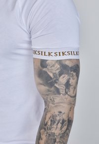 Balta prigludusi marškinėlė su tekstūruota medžiaga, ant rankovės yra kontrastinga auksinė juosta, kurioje pavadinimas "SikSilk."