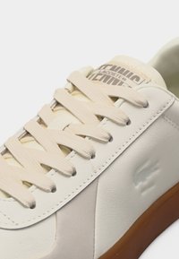 Λευκό δερμάτινο αθλητικό παπούτσι με μπεζ κορδόνια, ανάγλυφο λογότυπο κροκοδείλου στο πλάι και σόλα από καουτσούκ καφέ χρώματος.