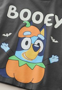 Szara koszulka z postacią psa z kreskówki w stroju dyni, z wyraźnym napisem "BOOEY" oraz małymi grafikami nietoperzy w kolorze białym.