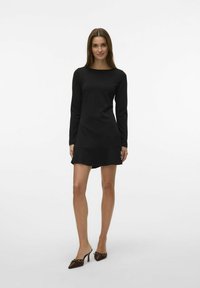 Vestido negro de manga larga con escote redondeado, dobladillo acampanado y textura suave. Combinado con mule de tacón marrones con acentos decorativos.