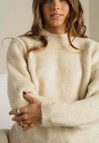 Cremefarvet sweater med ribbet rund halsudskæring og blødt, tekstureret stof. Har lange ærmer og let flagrende manchetter.