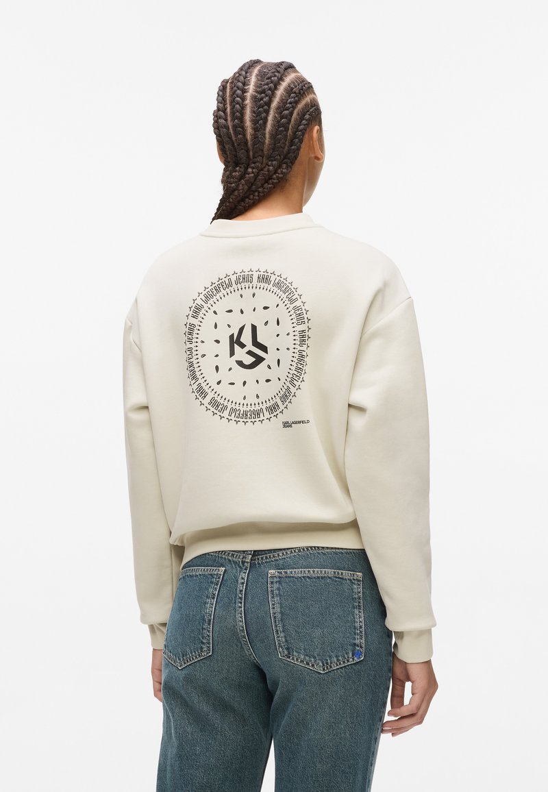 Bež hooded sweatshirt s krožnim grafičnim dizajnom na hrbtu, ki vsebuje besedilo in logo v črni barvi. V kombinaciji z modrimi jeans hlačami.