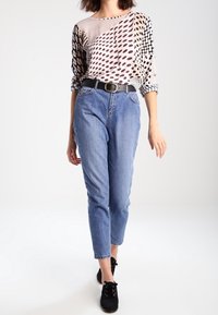 Top beige morbido con motivi in bianco e nero. Jeans in denim blu a vita alta. Cintura nera e scarpe nere con lacci. Texture complessiva liscia.