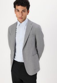 Blazer grigio chiaro con una finitura testurizzata, dotato di due tasche frontali e revers a punta, indossato sopra una camicia a righe blu e bianca.
